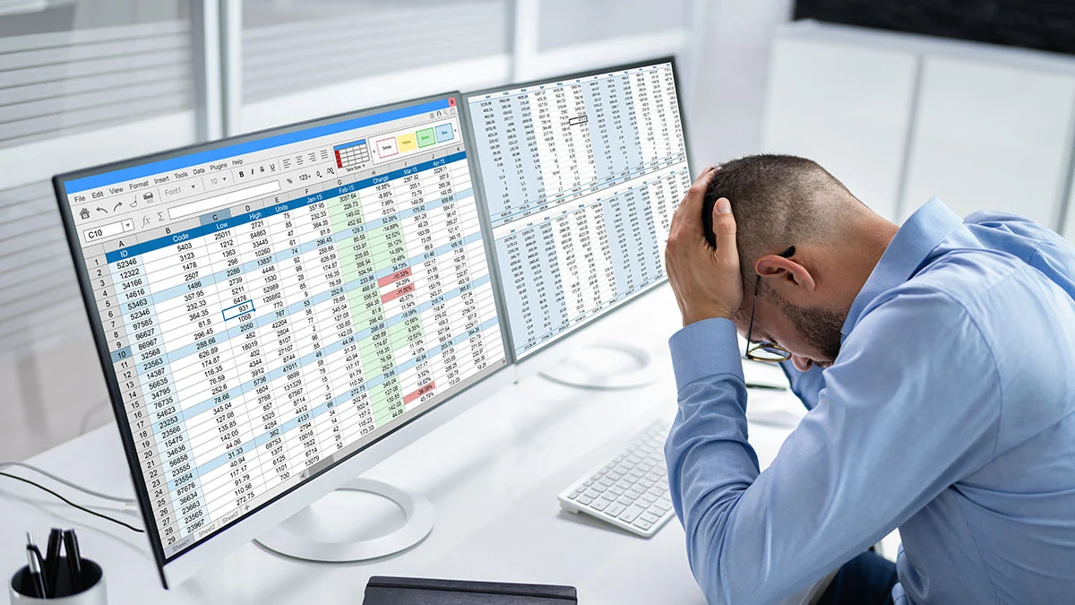 Seorang pria profesional terlihat stres di depan dua layar monitor berisi spreadsheet Excel penuh data angka dan tabel.