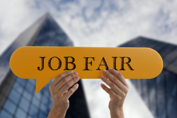 Job Fair: Hanya Sekadar Formalitas? Dua tangan memegang papan kuning bertuliskan "JOB FAIR" dengan latar gedung pencakar langit.