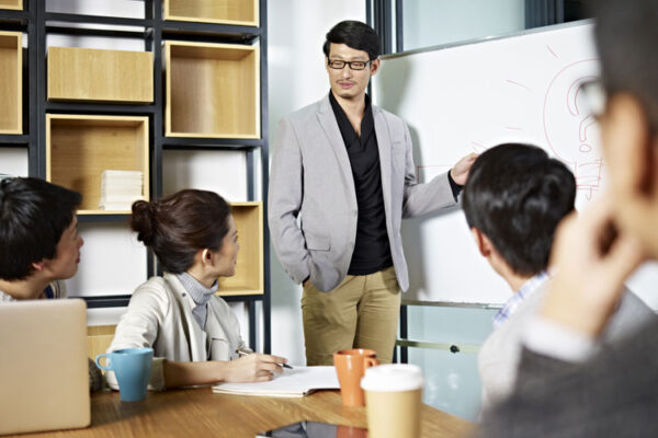 lustrasi seorang Product Manager memimpin tim, berdiskusi dengan laptop dan papan strategi produk.