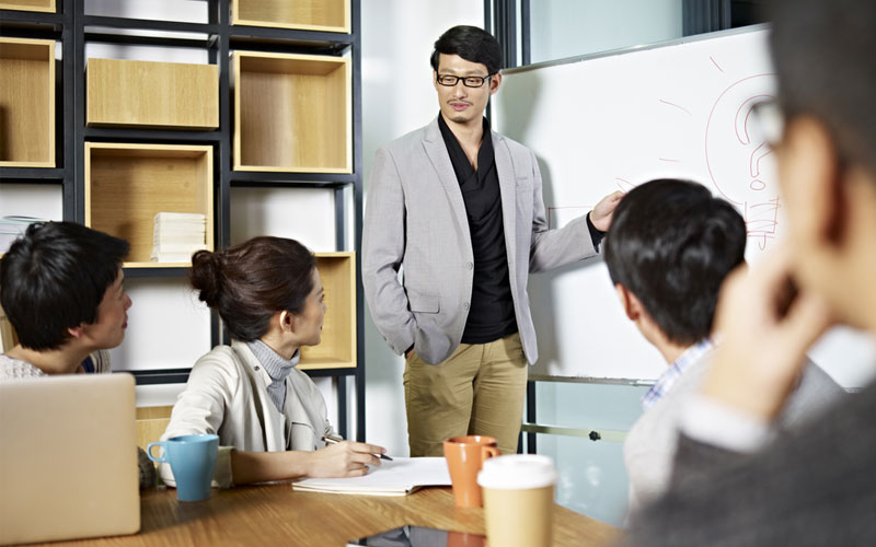 lustrasi seorang Product Manager memimpin tim, berdiskusi dengan laptop dan papan strategi produk.