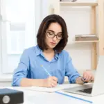 ilustrasi fresh graduate perempuan berbaju biru sedang mengerjakan dummy project untuk portofolio di depan laptop sambil menulis ide dan perencanaan proyek.