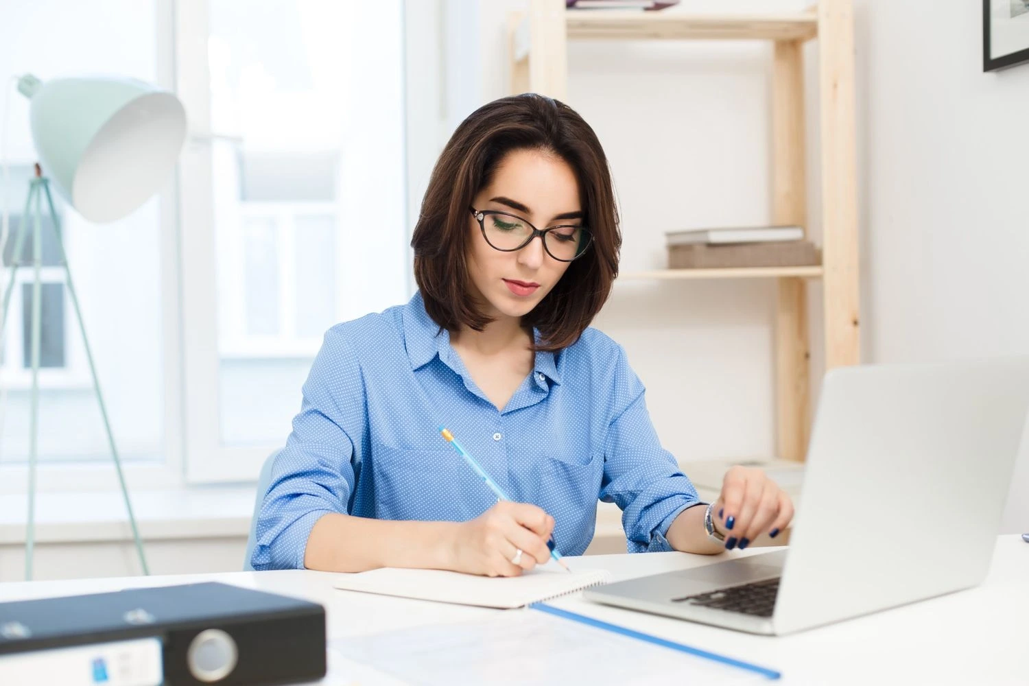 ilustrasi fresh graduate perempuan berbaju biru sedang mengerjakan dummy project untuk portofolio di depan laptop sambil menulis ide dan perencanaan proyek.