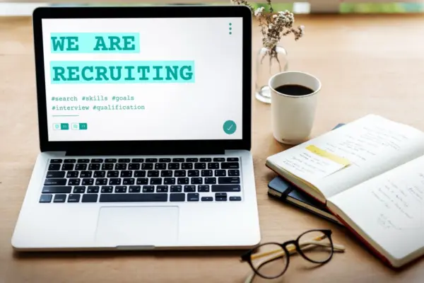 Laptop dengan tulisan “We Are Recruiting” di layar, ditemani buku catatan, kopi, dan kacamata di meja kerja sebagai illustrasi bolehkah melamar lagi diperusahaan yang sama