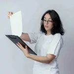 Seorang mahasiswa berkacamata dan berbaju putih sedang melihat contoh proposal magang mandiri untuk dijadikan referensi.